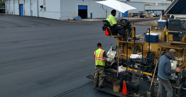 Three workers on an asphalt laying machine paving a new parking lot in front of a white building